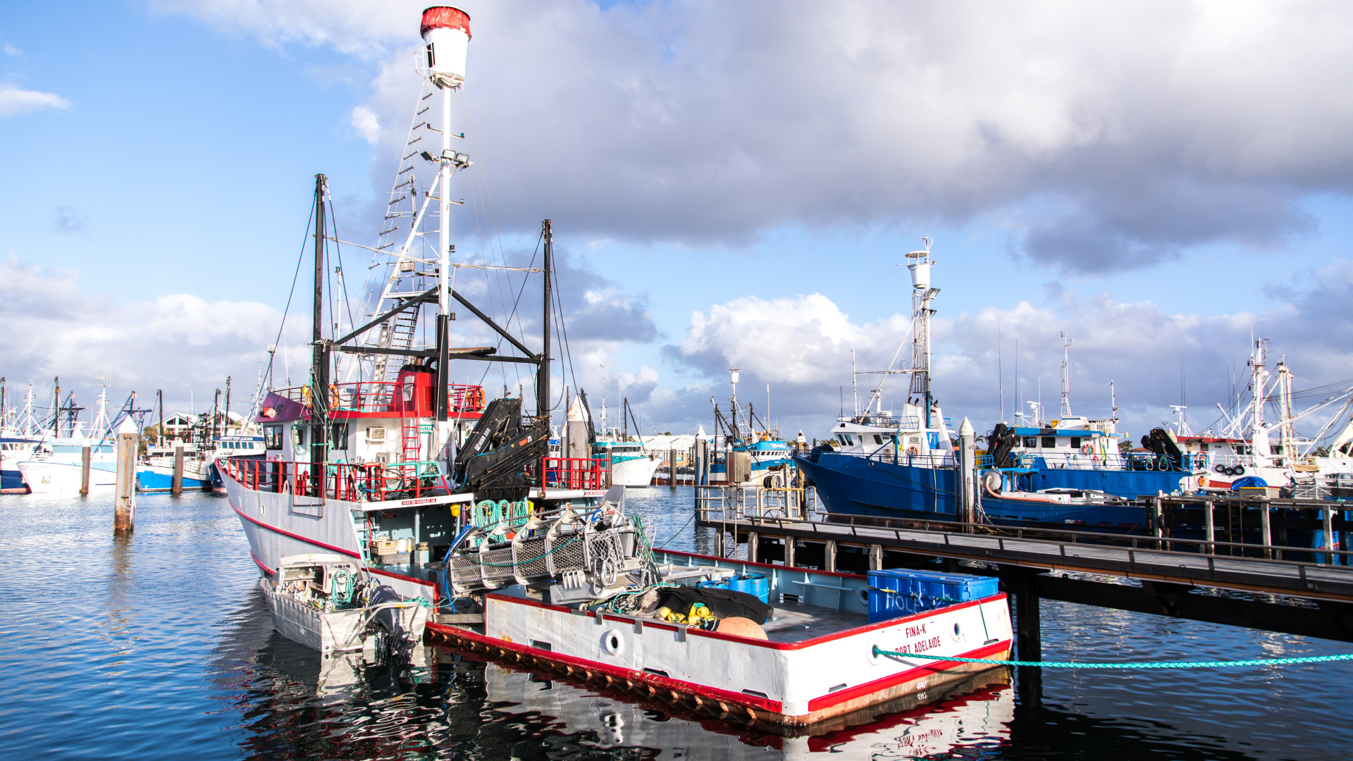 A cluster of commercial fishing boats, including a prominent red-and-white vessel with equipment on deck, are docked at a bustling marina. The scene is brightly lit by afternoon sun, with blue skies, scattered clouds, and reflections shimmering on the calm water.