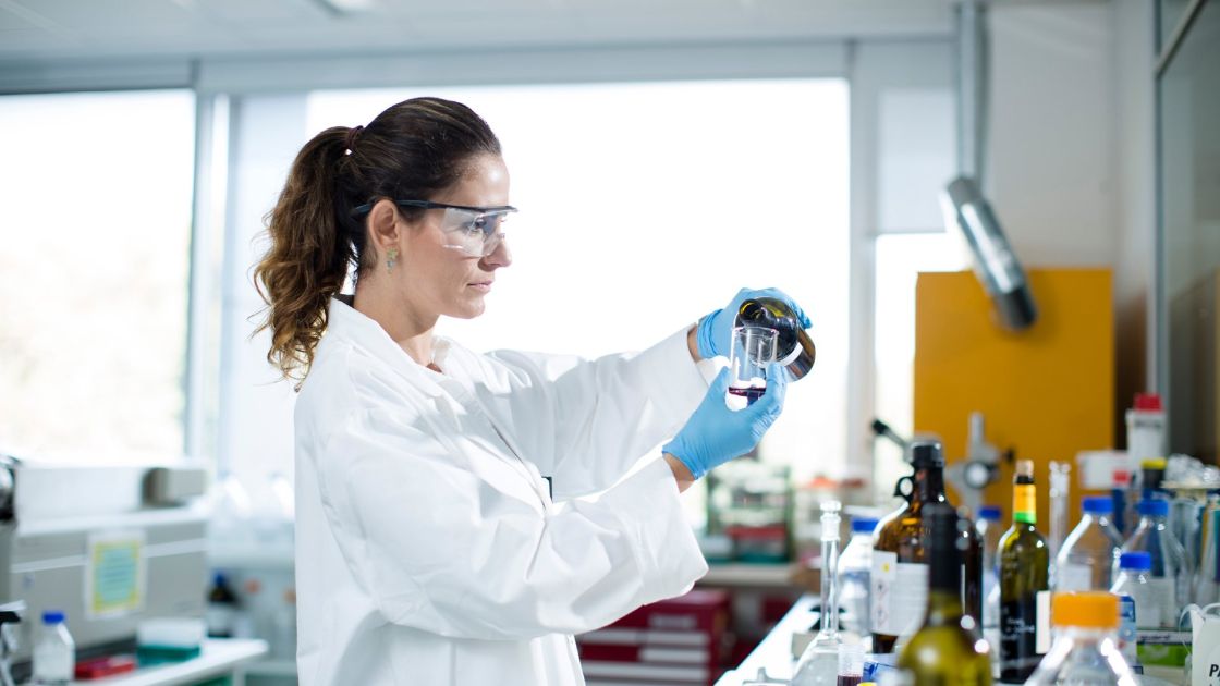A woman wearing safety glasses and a lab coat pouring wine into a beaker in a research laboratory.