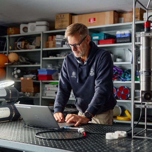 A man standing at a bench, working at a laptop with shelves of equipment in the background.