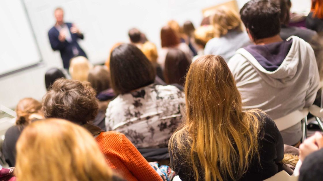 The backs of people's heads in a crowd listening to a person who is speaking.