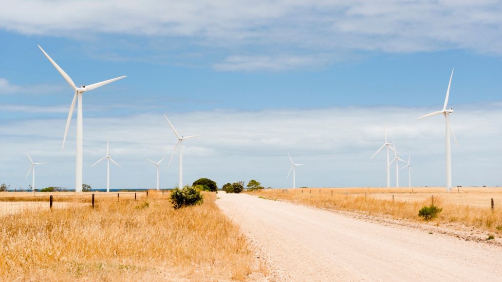 Wind turbines stand in a line along both sides of a dirt road that is stretching towards the horizon. 