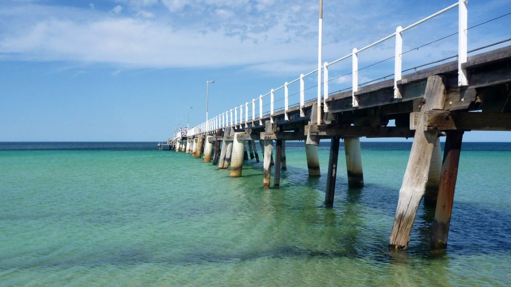 A wooden jetty stretches into a pale ocean with deeper blue water in the distance. White clouds spread across the blue sky.