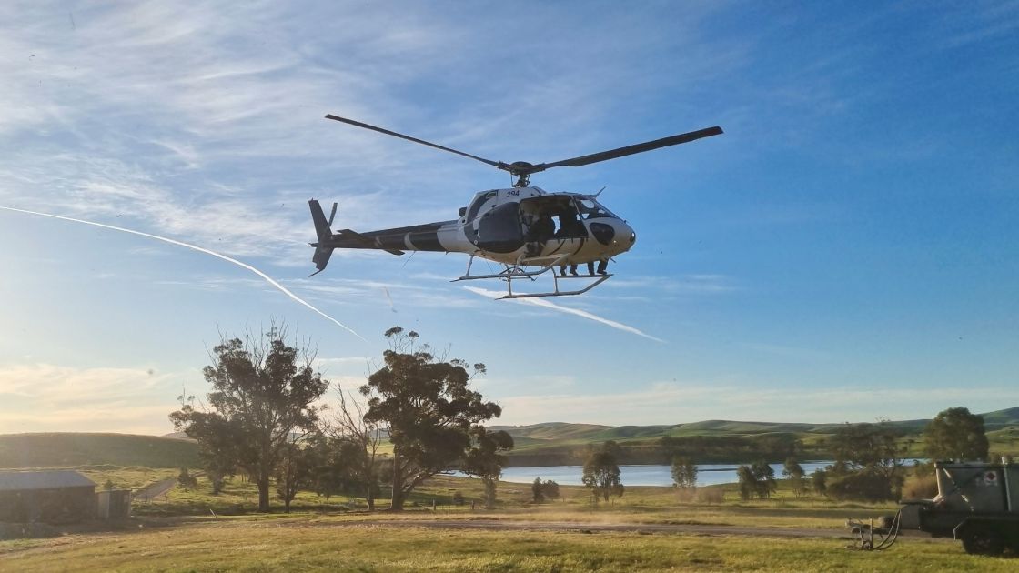 A helicopter is low in the sky with a farm property in the background.