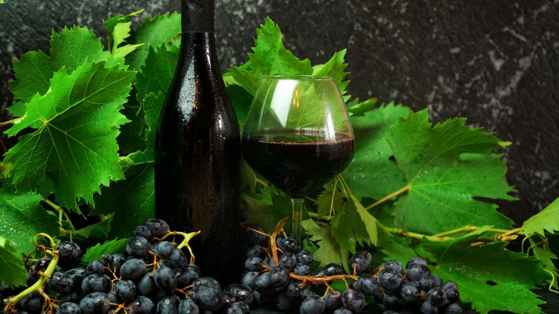A bottle of red wine next to a glass with wine in it, sitting among grapes and vine leaves.