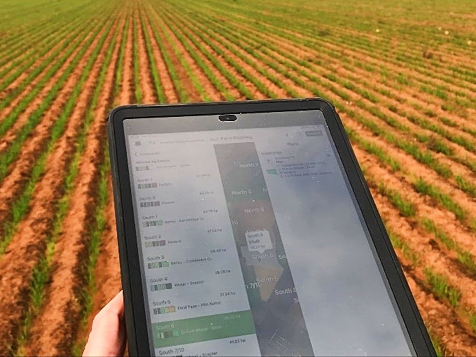 A mobile tablet displaying the Agworld platform with rows of crops in the background.