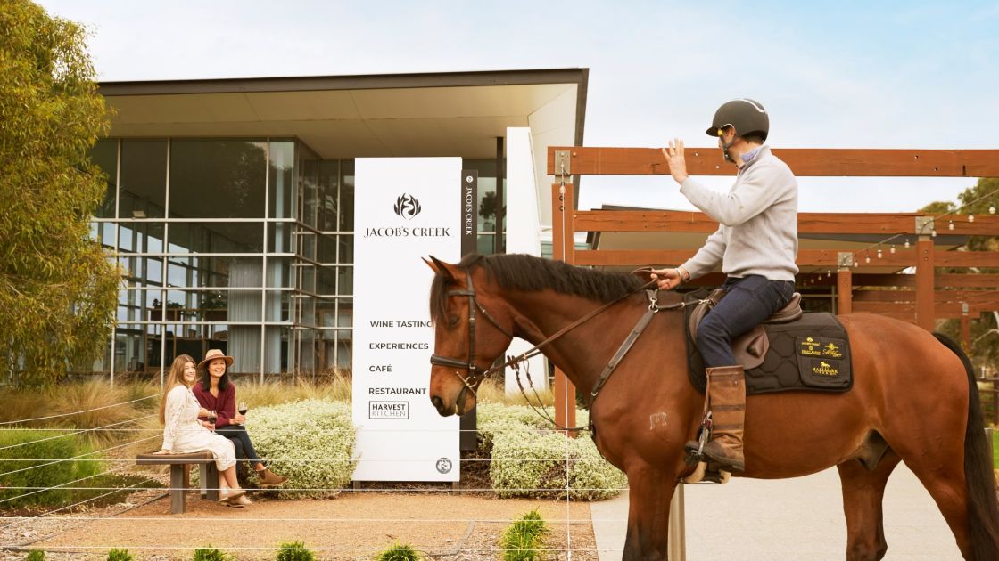 A man on a horse waves at 2 women sitting on a bench with glasses of wine outside of a building.