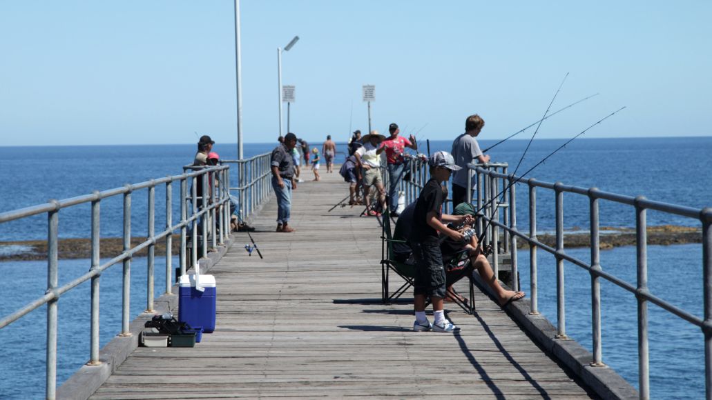 People of all ages fishing off a wooden jetty on a calm, sunny day.